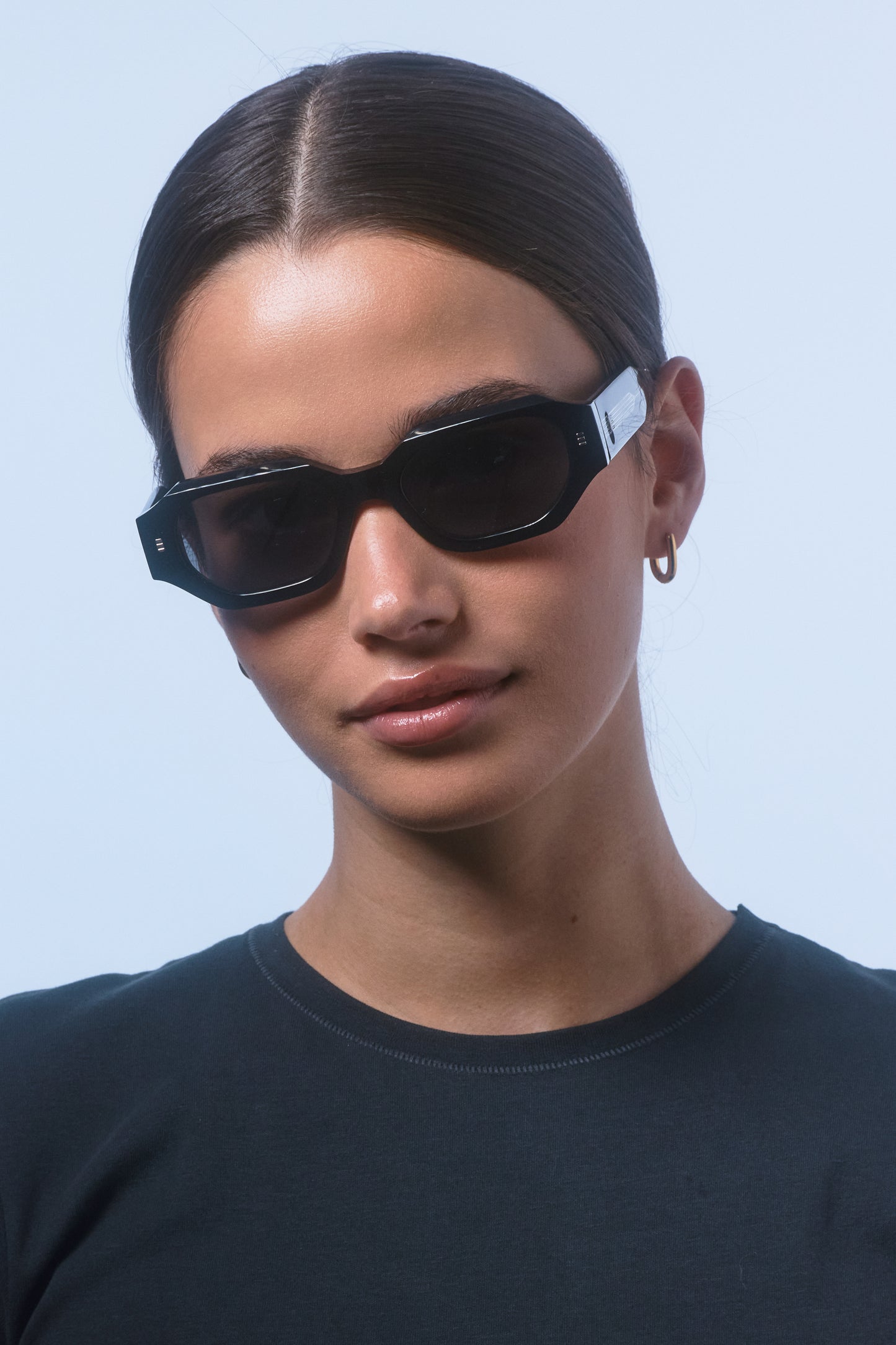 Woman wearing Isle of Eden Blake black hexagonal sunglasses, front-facing portrait against a light blue background