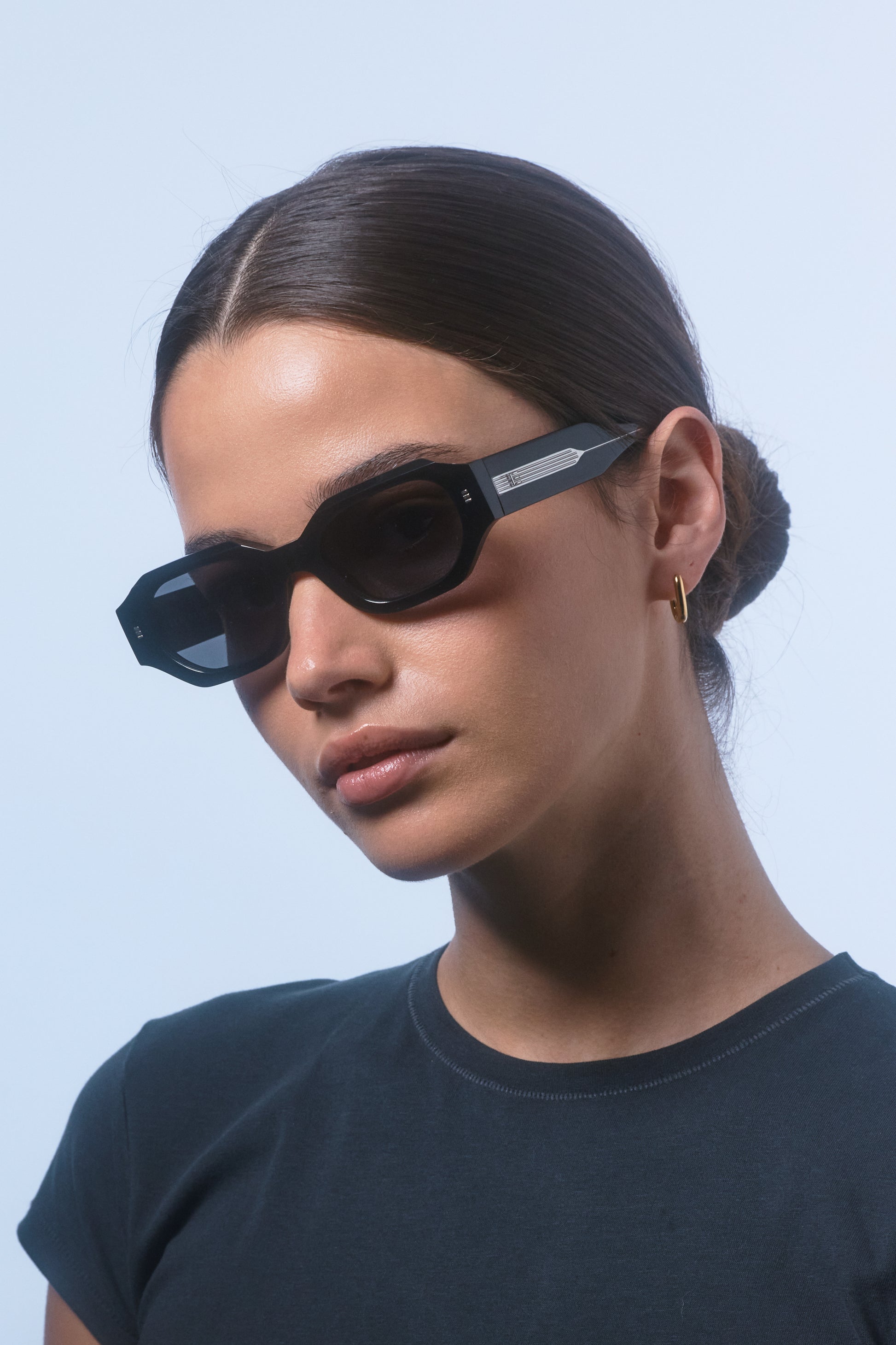 Woman wearing Isle of Eden Blake black hexagonal sunglasses, angled portrait against a light blue background