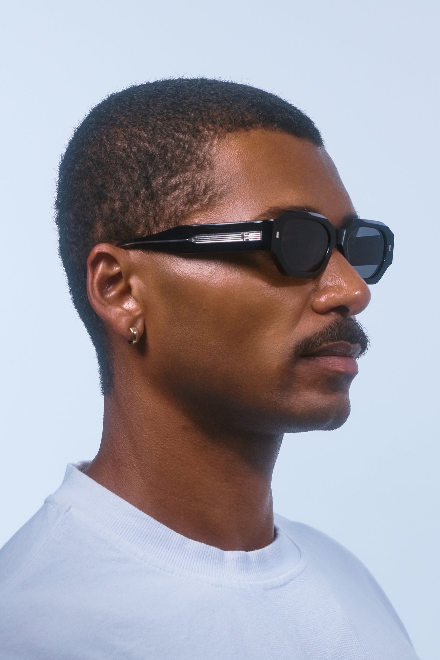 Man wearing Isle of Eden Blake Black Wire hexagonal sunglasses, side profile portrait against a light blue background