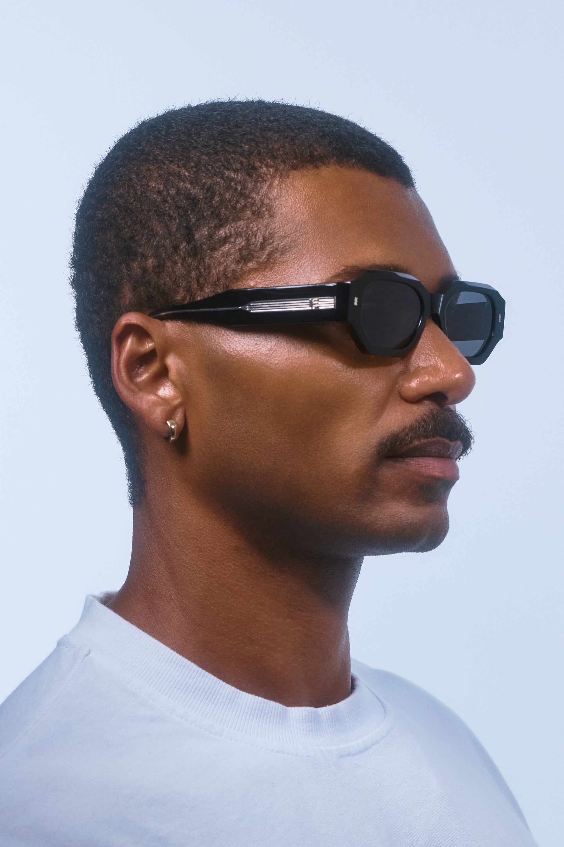 Man wearing Isle of Eden Blake Black Wire hexagonal sunglasses, side profile portrait against a light blue background