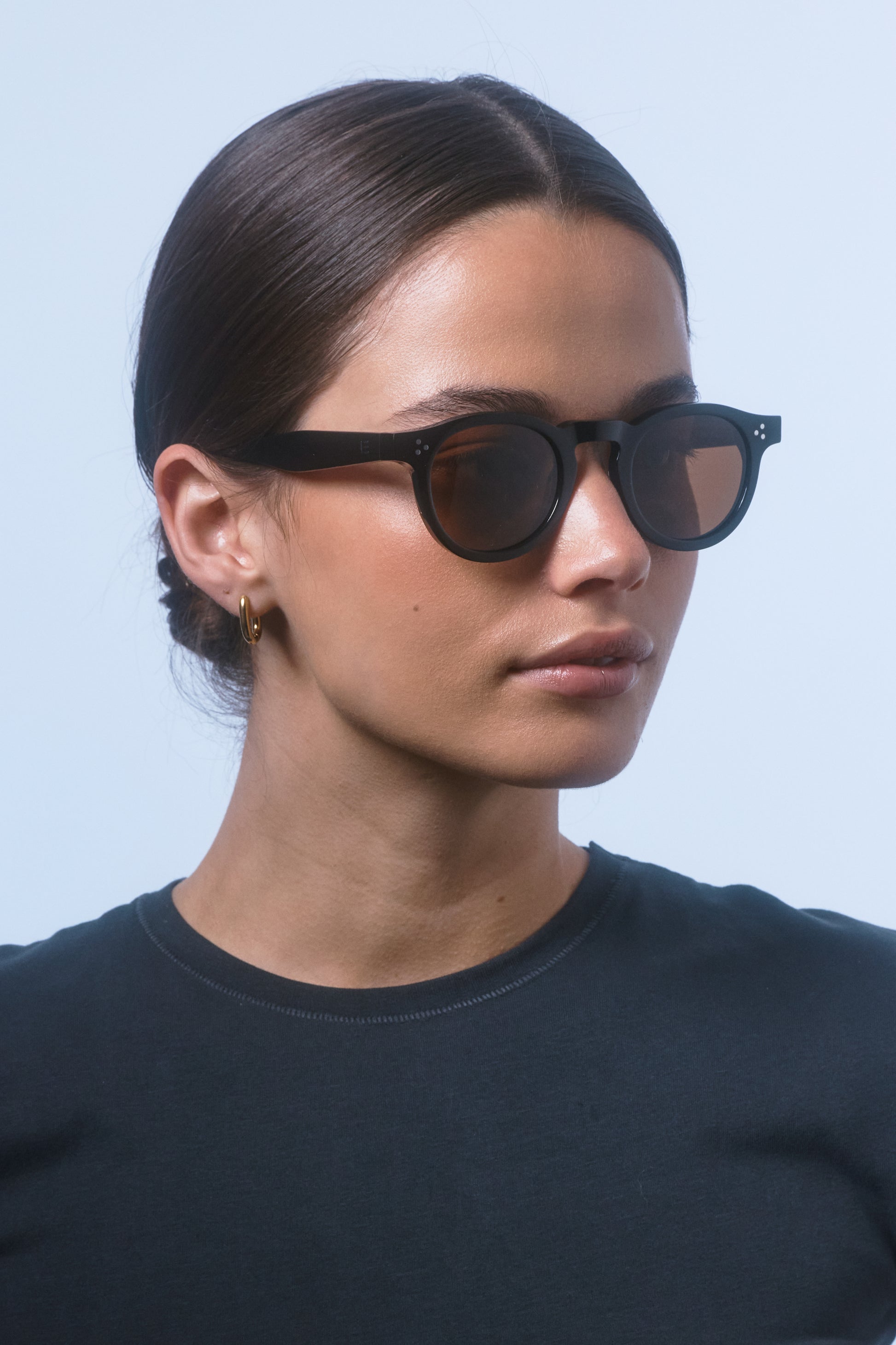 Female model in a black shirt wearing Isle of Eden Drew Black Prescription round bio acetate sunglasses, looking to the side.