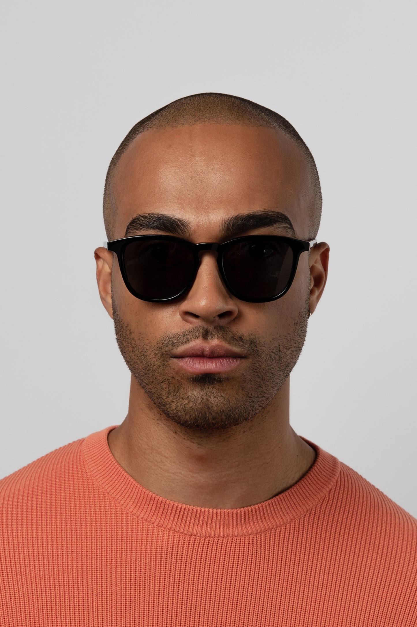 Man wearing Isle of Eden Louis-Philippe black sunglasses, front-facing portrait against a grey background